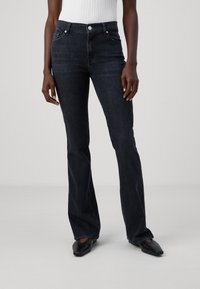 Jean évasé en denim foncé avec une taille ajustée, comportant cinq poches et des détails de couture subtils ; porté avec des chaussures plates noires.