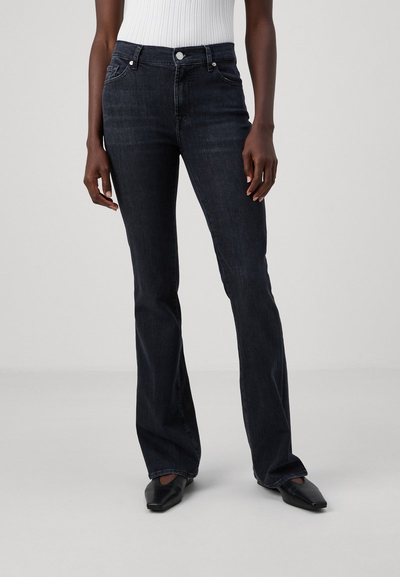 Jean évasé en denim foncé avec une taille ajustée, comportant cinq poches et des détails de couture subtils ; porté avec des chaussures plates noires.