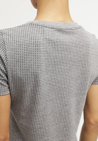 Pohled zezadu na osobu, která má na sobě šedou košili s krátkým rukávem, pokrytou malým černým čtvercovým vzorem na bílém pozadí.