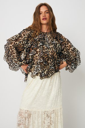 Γυναίκα που φοράει μπλούζα με βολάν και λεοπάρ σχέδιο από διαφανές ύφασμα και μακριά λευκή φούστα από δαντέλα, στέκεται με τα χέρια στη μέση.