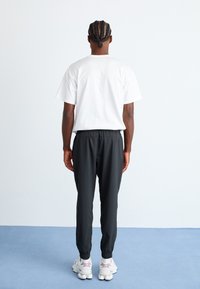 Person med fletninger står foran en hvid væg iført en hvid T-shirt, sorte joggers og hvide sneakers på et blåt gulv.
