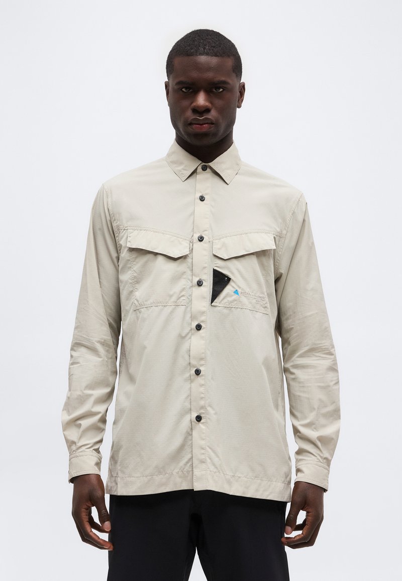 Άνδρας που φοράει μια μπεζ πουκάμισο με κουμπιά και δύο τσέπες στο στήθος, η μία με μαύρη και μπλε τριγωνική λεπτομέρεια, στέκεται μπροστά σε λευκό φόντο.