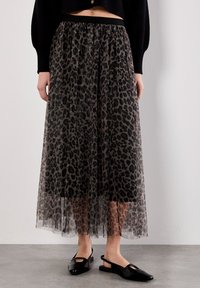 Eine Frau trägt einen langen, durchsichtigen Rock mit Leopardenmuster über einem schwarzen Unterrock, kombiniert mit schwarzen Slingback-Flats und einem schwarzen, kurz geschnittenen Pullover.