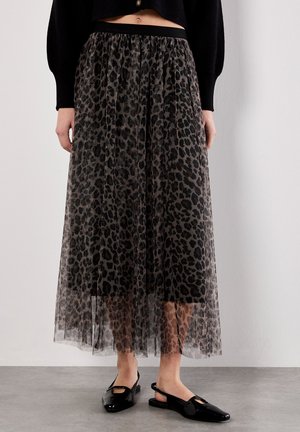 Femme portant une longue jupe en voile imprimé léopard par-dessus une jupe noire, associée à des ballerines noires à talon et à un pull noir court.