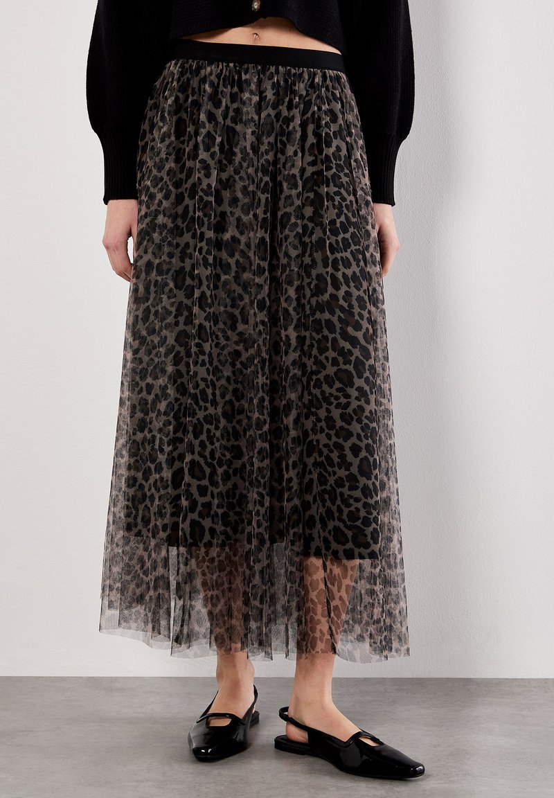Eine Frau trägt einen langen, durchsichtigen Rock mit Leopardenmuster über einem schwarzen Unterrock, kombiniert mit schwarzen Slingback-Flats und einem schwarzen, kurz geschnittenen Pullover.
