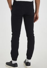 Pantalon de jogging noir en coton avec une coupe fuselée, des poignets élastiqués, deux poches arrière et une texture lisse. Porté avec des baskets noires.