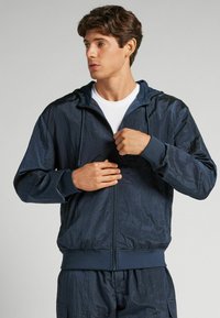 Mörkblå vindjacka med dragkedja fram, huva och ribbade ärmslut. Tillverkad av ett lätt, skrynkligt material med en texturerad yta.