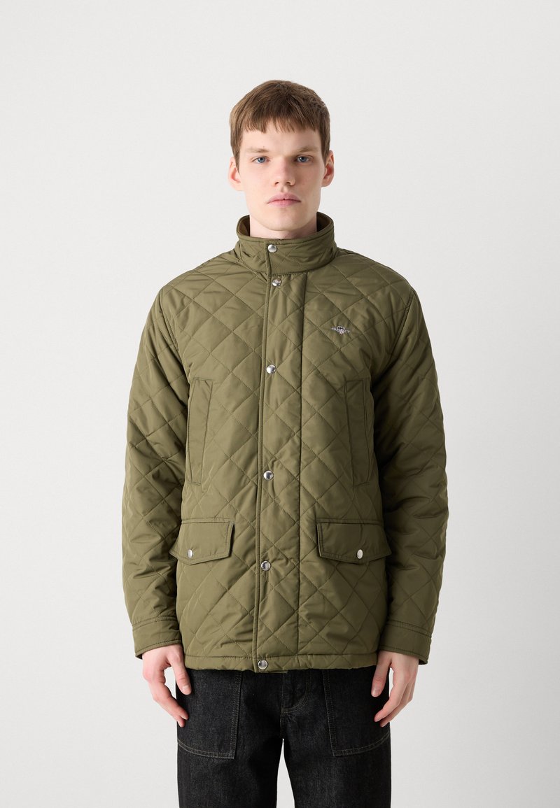 Zielona oliwkowa kurtka pikowana z wysokim kołnierzem, zatrzaskami, dwiema przednimi kieszeniami i subtelnym logo na klatce piersiowej. Gładka tekstura.