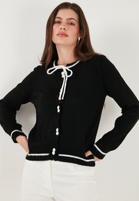 Črn pleteni kardigan z belimi detajli, ki ima zanko za zapiranje pri ovratniku, dolge rokave in bele gumbe spredaj.