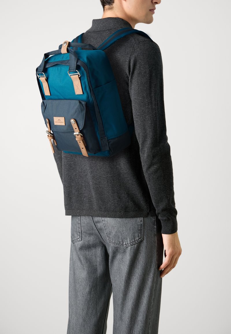 Tyrkysový batoh s hnědými koženými akcenty a nastavitelými popruhy, obdélníkového tvaru s přední kapsou, vyrobený z odolné tkaniny.