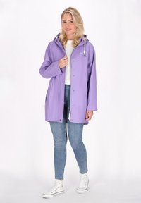 Ljuslila vattentät jacka med huva, tryckknappar och dragsko. Bärs över en vit topp och blå jeans, kompletterad med vita sneakers.