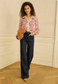 Blúzka s kvetinovým vzorom, dlhé rukávy, výstrih do V a čipkový lem, v kombinácii s tmavomodrými džínsami s širokými nohavicami a svetlohnedou mäkkou kabelkou.