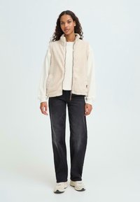 Gilet in fleece beige con colletto alto, zip frontale e due tasche laterali, indossato sopra una camicia bianca a maniche lunghe e abbinato a pantaloni neri.