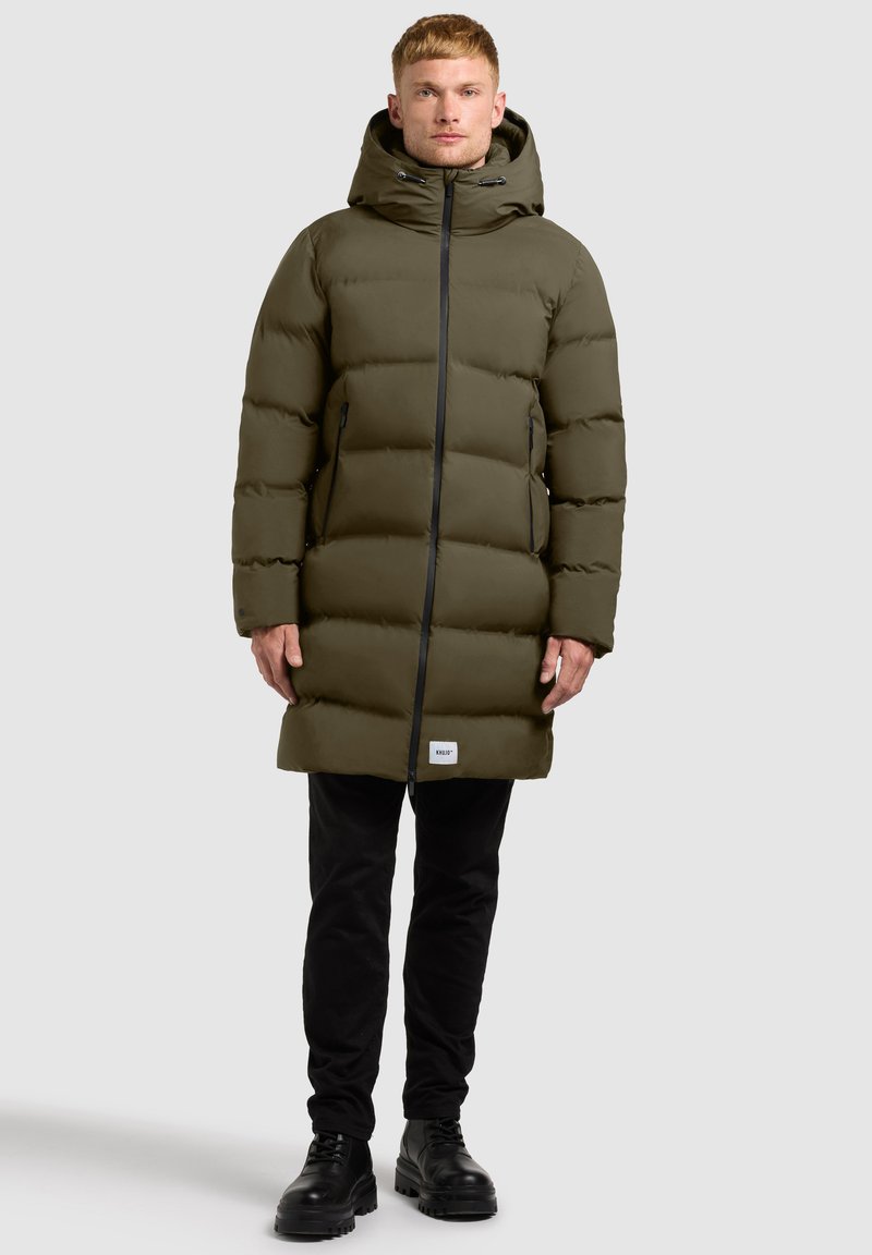 Geacă puffer verde olive cu glugă, având un fermoar frontal, accente negre și două buzunare laterale cu fermoar. Pur tată cu pantaloni negri și botine.
