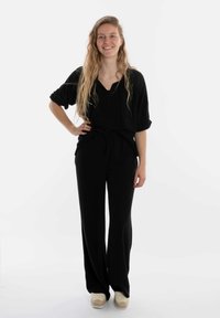 Zwarte, losvallende jumpsuit met een v-halslijn en opgerolde mouwen, vastgemaakt rond de taille. In combinatie met lichtgekleurde plateauschoenen.