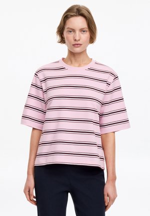Personne portant un t-shirt rose à manches courtes, ample, avec des rayures noires horizontales et un pantalon noir, debout devant un fond blanc uni.