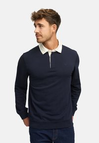 Granatowy sweter z białym kołnierzykiem i rozpięciem na dwa guziki. Wykonany z miękkiego materiału, ma ściągacze na mankietach i u dołu, a także subtelne logo na klatce piersiowej.