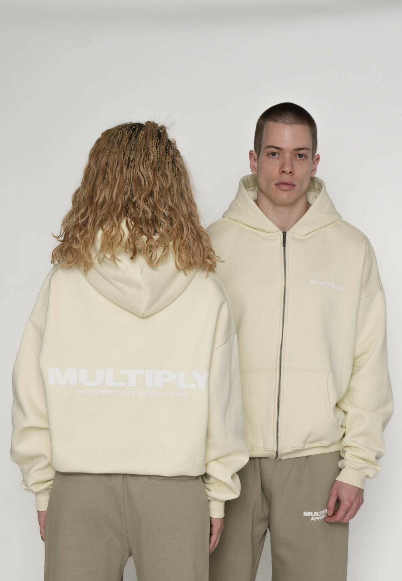 Kremfarvede hættetrøjer med lynlås og "MULTIPLY"-logo i hvid. Afslappet pasform med ribbet kant ved ærmerne. Båret med matchende lysebrune bukser.