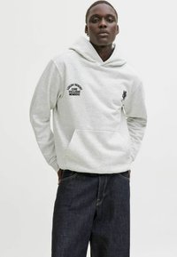 Jasnoszara bluza z kapturem z kieszenią kangurzą, z czarnym haftowanym napisem i małą grafiką. Połączona z ciemnoniebieskimi dżinsami.