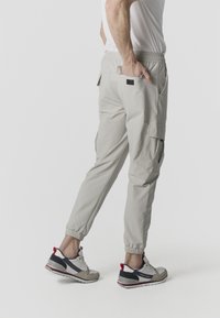 Hombre con pantalones jogger cargo beige y zapatillas blancas con detalles en rojo y negro, de pie con una mano en el bolsillo trasero.