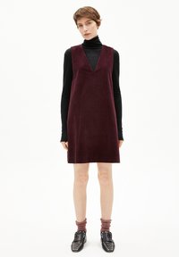 Bordeauxfarbene Cordkleid mit V-Ausschnitt, kombiniert mit einem schwarzen Rollkragenpullover. Das Model trägt schwarze Schuhe mit silbernen Akzenten und gerippte Socken.