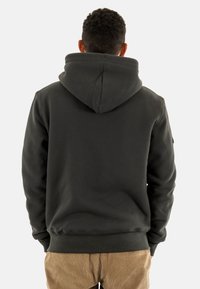Sweatshirt à capuche noire en matériau doux, avec poche kangourou et poignets côtelés, porté avec un pantalon en velours côtelé beige. Vue arrière montrée.