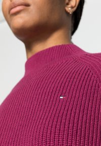 Trui in ribbel tricot, diep magenta kleur, ontwerp met hoge nek. Bevat een subtiel logo in rood, wit en blauw aan de linkerkant. Zachte textuur.