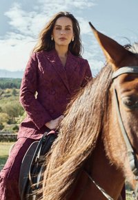 Burgundy textured double-breasted blazer with a matching skirt. Model sits on a horse, with long hair cascading over the saddle.