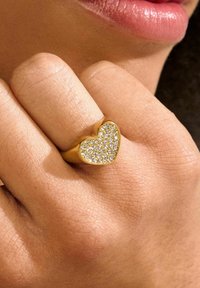 Anillo dorado en forma de corazón engastado con pequeñas piedras transparentes, con una banda lisa y un acabado pulido, posado sobre una mano con textura de piel natural.