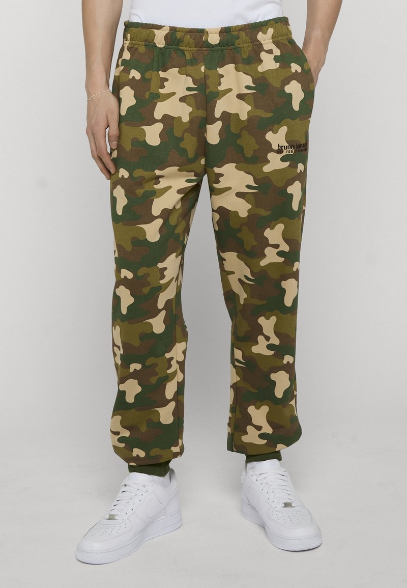 Homme portant un pantalon camouflage vert et marron à taille élastique et des baskets blanches, les mains dans les poches, sur un fond gris uni.