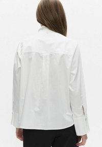 Biała koszula z długimi rękawami i rozciętymi mankietami, z zapięciem na guziki oraz panelem yoke z tyłu. Gładki materiał.