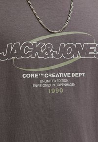 Kratak prikaz sive tkanine s bijelim i zelenim tekstom: "JACK & JONES," "CORE™ CREATIVE DEPT. UNLIMITED EDITION. ENVISIONED IN COPENHAGEN 1990," i srebrnim lancem.