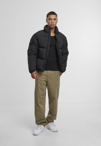 Svart pufferjakke med høy krage, quiltet tekstur, brukt over en svart skjorte, kombinert med khaki bukser og hvite joggesko.