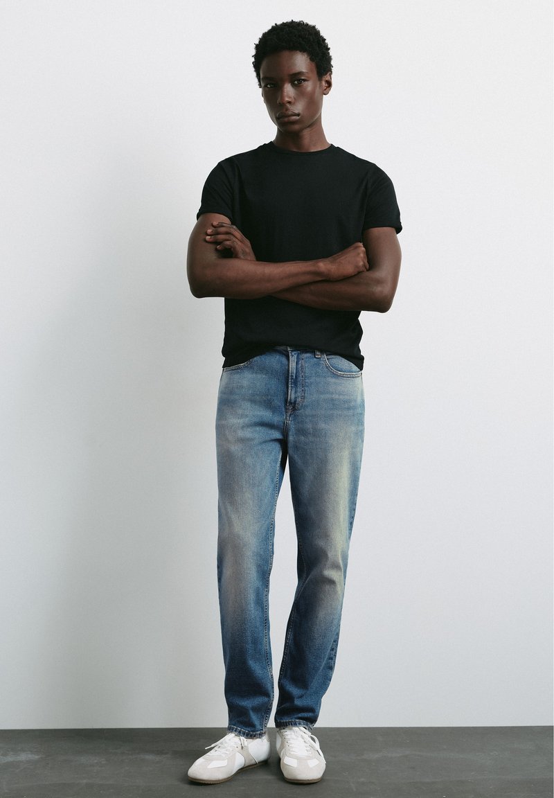 Jeune homme aux cheveux courts et bouclés, bras croisés, portant un t-shirt noir, un jean bleu clair et des baskets blanches devant un mur uni.