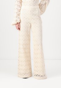 Pantaloni in pizzo color crema con motivo a zigzag, gamba larga e tessuto testurizzato. Presentano una vita a metà e un design fluido.