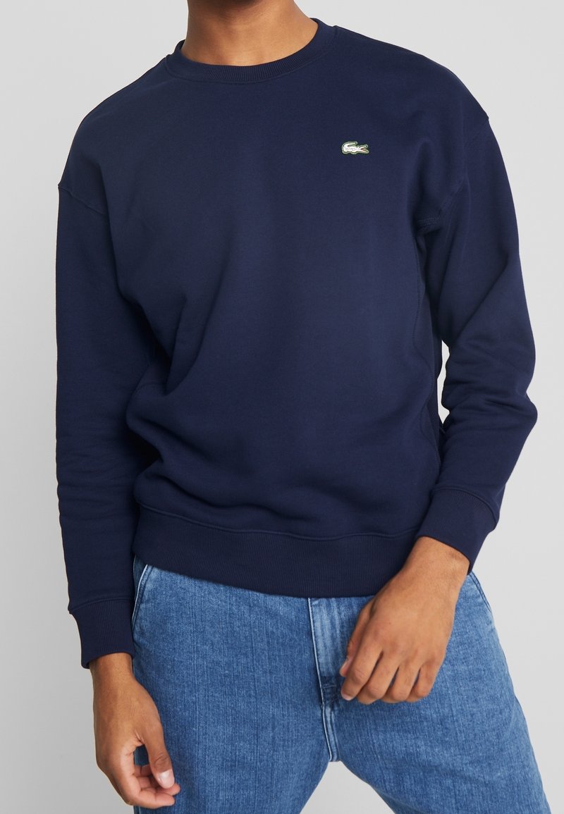 Personne portant un sweat-shirt bleu marine à manches longues avec un petit logo crocodile vert sur la poitrine et un jean bleu.