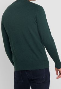 Man in een donkergroene lange mouwen trui en donkerblauwe jeans, gezien van achteren tegen een effen lichtgrijze achtergrond.