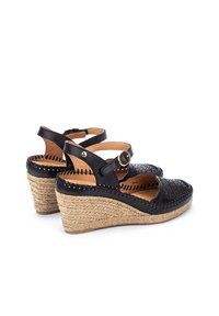 Sandalias de cuña de cuero negro con diseño perforado, tirante en el tobillo con hebilla y tacón de cuña envuelto en yute, mostrando una textura natural.