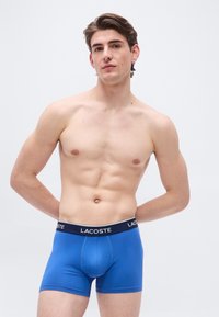 Άνδρας χωρίς μπλούζα με κοντά καστανά μαλλιά, φορώντας μπλε εσώρουχα Lacoste boxer, που στέκεται με τα χέρια πίσω από την πλάτη του μπροστά σε απλό λευκό φόντο.