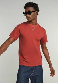 T-shirt rossa a girocollo con maniche corte, caratterizzata da un logo sottile. Modello che indossa occhiali da sole neri, posato su uno sfondo chiaro.