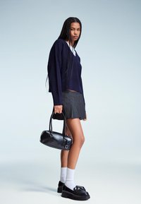 Ženska z dolgimi kitami, oblečena v mornarsko pulover, karirasto plisirano krilo, bele nogavice, črne platforme in kupčka, drži črno usnjeno torbico.