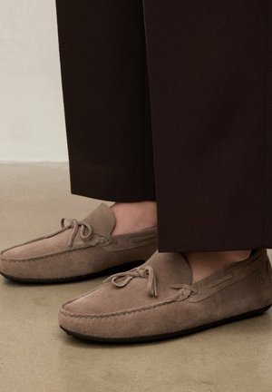 Brune ruskind loafers med sløjfedetaljer båret med mørkebrune, vidde bukser på en glat beige gulv.