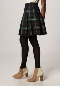 Falda en línea A de cuadros en colores oscuros con líneas verdes, amarillas y rojas. Se lleva con medias negras y botas de tobillo beige con tacón cuadrado.