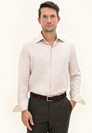 Uomo con capelli castani corti che indossa una camicia beige a maniche lunghe abbottonata, pantaloni scuri e cintura marrone, in piedi con una mano in tasca.