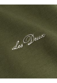 Tessuto verde oliva con texture visibile e il testo ricamato "Les Deux" in bianco, caratterizzato da un dettaglio in font corsivo.