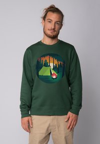 Groene sweater met een grafisch ontwerp van een tent en een kampvuur binnen een cirkel, met een achtergrond van een zonsondergang en een bos.