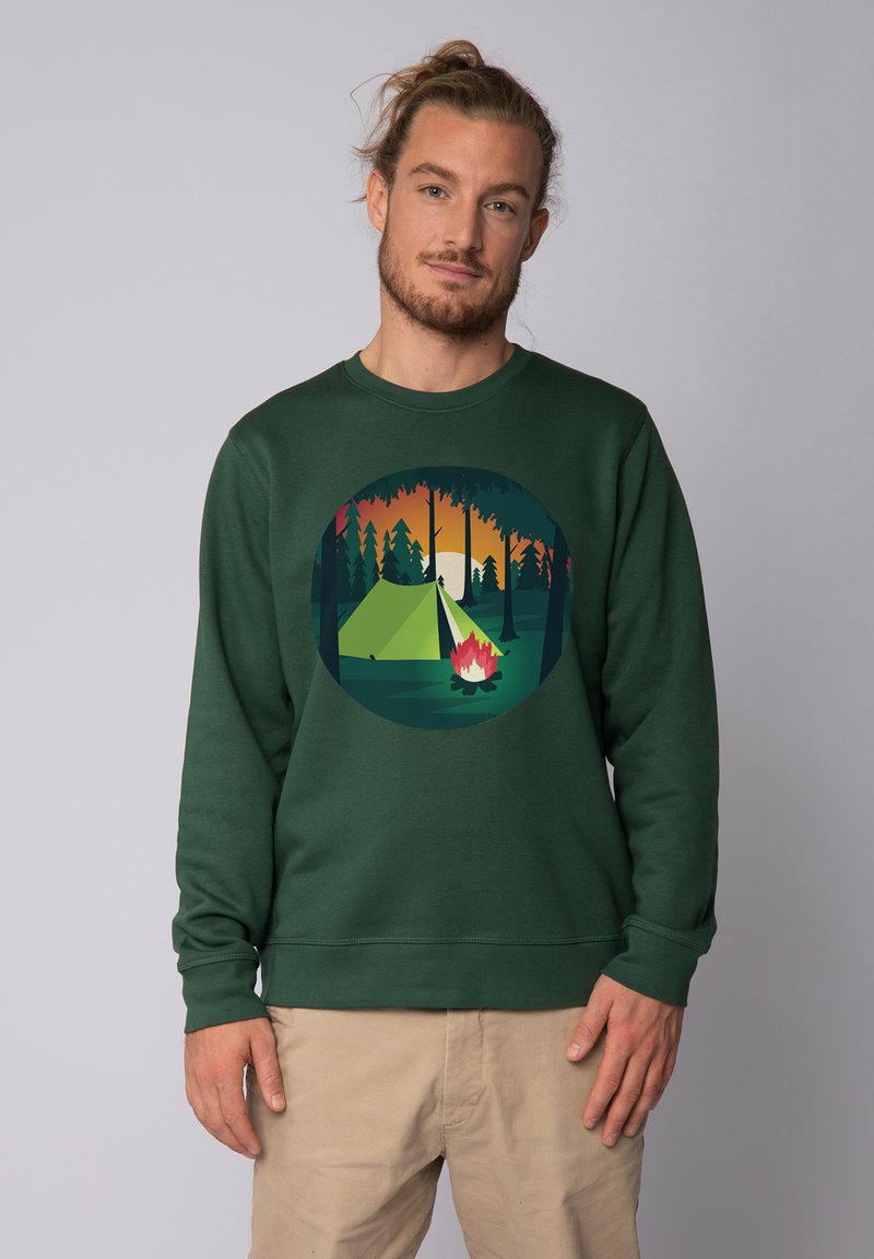 Groene sweater met een grafisch ontwerp van een tent en een kampvuur binnen een cirkel, met een achtergrond van een zonsondergang en een bos.