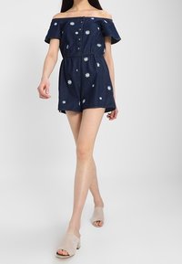 Woman wearing off-shoulder navy blue romper with white daisy prints and button details, paired with beige open-toe block heels.