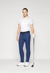 Abbigliamento da golf composto da una polo bianca e pantaloni blu navy con una cintura nera. Il modello tiene una mazza da golf e indossa scarpe bianche.