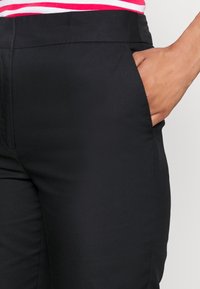 Pantaloni neri in tessuto liscio, dotati di tasche laterali e di un design aderente, mostrati in vita con una maglietta a righe parzialmente visibile.